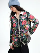 Camisa de corte relajado con estampado floral vibrante sobre fondo negro. Presenta cuello clásico, cierre frontal con botones y puños en contraste. El diseño destaca por sus terminaciones redondeadas y delicadas.