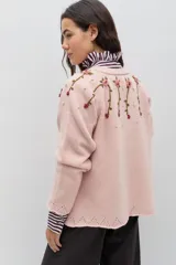 Saco de punto color rosa, con cuello redondo, cierre frontal con botones forrados en crochet y mangas largas. Presenta un delicado bordado floral artesanal que cae desde los hombros y detalles de calados en los puños y el ruedo.