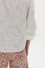 Camisa blanca de algodón, de corte clásico y atemporal, con cuello mao, mangas abullonadas y detalle de volados en la botonera frontal oculta.