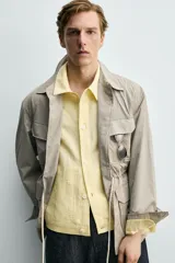 Parka relaxed fit color gris, confeccionada en tejido técnico ligero, con cuello solapa, manga larga con puño abotonado, cuatro bolsillos de plastrón con solapa en el frente, cintura ajustable con cordón y cierre frontal de botones.