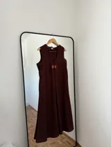 Vestido largo sin mangas, color marrón oscuro, con escote redondo y corte amplio. Presenta bolsillos laterales.