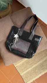 Tote bag de ecocuero negro con textura tipo cocodrilo, cuenta con un bolsillo frontal de cuero liso en tono marrón oscuro. Incluye un charm colgante con cadena metálica y una estrella decorativa. Posee doble asa de hombro y cierre superior.