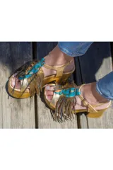Sandalias de cuero dorado con plataforma de madera, taco alto, flecos prominentes y detalle frontal de cuero pintado a mano en tonos turquesa y cobre.