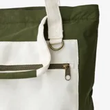 Bolso tote verde militar confeccionado en nylon resistente al agua, con un bolsillo frontal grande en color crudo que incluye cierre horizontal y una anilla metálica. Cuenta con asas largas de hombro en color crudo y una manija superior corta.
