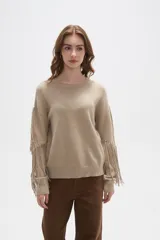 Sweater de punto color beige, con cuello redondo, corte holgado y diseño de flecos largos en las mangas.