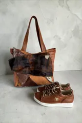 Cartera tipo tote de cuero en tono marrón, combinada con apliques de pelo vacuno animal print, detalles metalizados y dije colgante triangular.