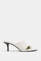 Sandalia tipo mule de tacón alto fino de madera, confeccionada en piel color blanco marfil. Presenta una tira asimétrica con separador para el dedo y punta redonda.