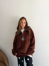 Buzo de corderito color marrón, con cuello alto y cierre frontal parcial. Presenta un diseño holgado con mangas largas y un parche decorativo en el frente.