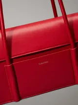 Cartera de hombro Calvin Klein estructurada, color rojo vibrante, con solapa frontal y bolsillo exterior con el logo grabado en metal plateado. Posee asas tubulares superiores y una correa ajustable para llevar cruzada.