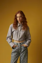 Camisa corta a rayas verticales grises y blancas, con cuello clásico, cierre frontal con botones y mangas largas con puños abotonados.