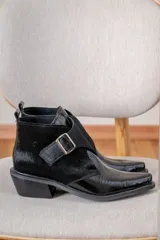 Botin de cuero negro con acabado brillante y detalle de solapa en cuero con textura de pelo, cierre con hebilla metálica lateral y punta fina.