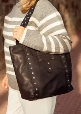 Bolso tipo tote bag de cuero negro con apliques metálicos plateados.