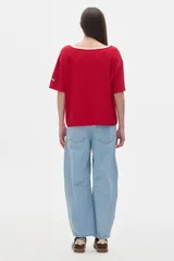 Remera oversized de manga corta en tejido rib blanco, con cuello redondo amplio ribeteado en negro. Presenta un pequeño parche rectangular con texto en la manga izquierda.