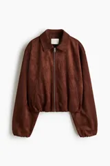 Campera corta marrón de tejido suave con terminación cepillada, cuello camisero, hombros caídos y cierre frontal. Cuenta con bolsillos laterales ribeteados y elástico revestido en puños y dobladillo.