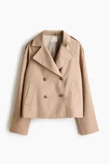 Gabardina corta color beige, de estilo cruzado con doble botonadura, solapas de muesca, bolsillos ribeteados y mangas raglán con charreteras en los hombros.