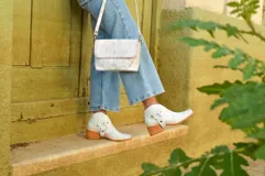 Cartera bandolera de cuero color blanco envejecido con pespuntes marrones. Tiene correa larga regulable y herrajes metálicos color bronce.