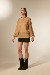 Blusa holgada color beige con textura plisada vertical y brillo sutil. Presenta cuello alto fruncido y mangas largas abullonadas con puños ajustados.