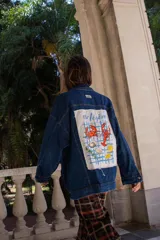 Campera de jean azul oscuro con corte oversize. Presenta un parche rectangular de tela blanca en la espalda con un estampado artístico de temática marina que incluye cangrejos, limones y la frase 'vie légère' sobre un fondo de cuadrícula azul.
