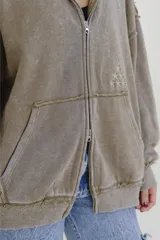 Canguro con cierre frontal, confeccionado en tela con efecto desgastado color beige. Presenta costuras expuestas, diseño holgado, capucha y un pequeño bordado de estrellas en el frente.