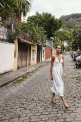 Vestido midi de color blanco con escote en pico y tirantes finos. Presenta un diseño con frunces en la cintura y detalle de lazada frontal.