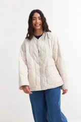 Campera acolchada de diseño liviano en color beige, con cuello redondo, cierre frontal con cremallera y dos bolsillos delanteros tipo parche. Presenta un patrón de costuras onduladas en toda la prenda.