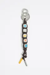 Llavero con mosquetón metálico plateado y cordón trenzado marrón. Incluye cinco dados metálicos con letras que forman la palabra "BOMBA" (B, O, M, B, A) con letras en colores azul y amarillo. Finaliza con un terminal metálico plateado y un detalle de cordón amarillo.