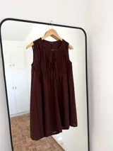 Vestido corto sin mangas color marrón oscuro, con escote redondo y detalle de pliegues verticales en el frente que terminan en un lazo ajustable en el busto.