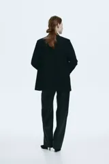 Blazer de sarga color negro con solapas de muesca, frente cruzado y cinturón fino ajustable con hebilla metálica. Presenta bolsillos delanteros ribeteados con solapa y diseño forrado.