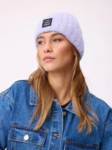 Gorro tipo beanie de punto acanalado en color lila, con parche rectangular negro en el frente que incluye la inscripción 'EAT THE WAVE'.