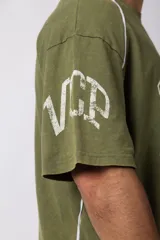 Remera de corte oversize color verde oliva con estampado frontal estilo universitario y detalles de líneas blancas en los hombros y laterales.