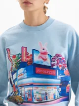 Buzo de cuello redondo y mangas largas, con silueta amplia y largo a la cadera. Presenta un estampado gráfico frontal de estilo urbano con temática de mercado y un conejo, parte de una colaboración artística.
