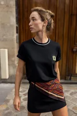 Camiseta negra de algodón, corte regular, con cuello redondo ribeteado en blanco y un bolsillo frontal con un broche metálico decorativo dorado.