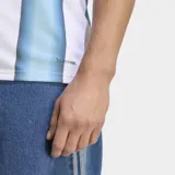 Camiseta de fútbol Adidas de la selección argentina, modelo Primera Equipación 26. Diseño a rayas verticales blancas y celestes con un efecto degradado en las franjas celestes. Presenta cuello redondo con ribete azul oscuro y tres franjas de Adidas en los hombros en azul oscuro. El escudo de la AFA y el logo de Adidas son de color dorado.