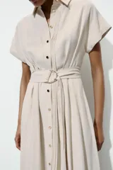 Vestido midi camisero de color beige, con cuello solapa, manga corta y cierre frontal con botones. Incluye un cinturón del mismo tejido con hebilla metálica.