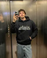 Canguro oversize de color negro con capucha y bolsillo frontal tipo canguro. Presenta un estampado gráfico central con estilo graffiti que incluye elementos urbanos y detalles en amarillo.