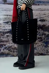Cartera tipo shopper de gran tamaño confeccionada en material efecto gamuza color rojo. Presenta correas reforzadas decoradas con tachas metálicas plateadas y un dije colgante en forma de corazón con detalles de tachas. Cuenta con una placa metálica pequeña con logo en la parte inferior frontal.