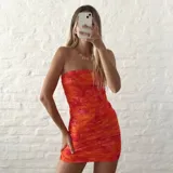 Vestido corto tipo strapless confeccionado en doble capa de microtul, con diseño ajustado y fruncido en toda la prenda. Presenta un estampado floral en tonos naranja y rojo.