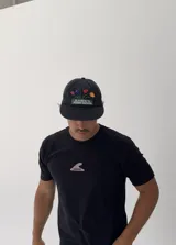 Gorro tipo gorra de béisbol color negro con visera curva. Presenta un bordado frontal de un macetero con flores de colores (rosa, azul, rojo y naranja) y el texto "OLIVER'S FLOWER GARDEN" en blanco sobre un fondo verde.