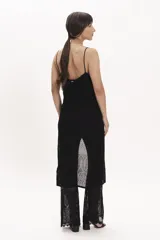 Pantalón de tejido de punto negro, corte ancho y tiro alto, con detalle de encaje calado en la parte inferior. Se lleva debajo de un vestido largo de encaje negro con tirantes finos y botones frontales.