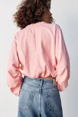 Blusa de manga larga con estampado de cuadros vichy en tonos rosado y blanco. Presenta cuello redondo con volante, cordón ajustable en el escote con apliques decorativos, y puños fruncidos con tiras para atar.