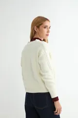 Cardigan de punto trenzado color crema con cuello redondo y cierre frontal de botones grandes en tono dorado. Presenta mangas largas con puños acanalados y un diseño de tejido con textura.