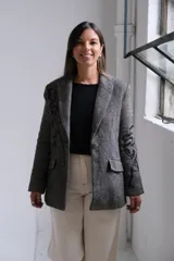 Blazer de corte sastrero en color gris con patrón de rayas diagonales y bordados negros en las mangas y solapas. Presenta cierre frontal con botón, bolsillos con solapa y estructura clásica.