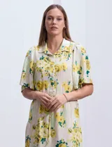 Vestido largo camisero con estampado floral amarillo y verde sobre fondo blanco. Tiene cuello camisero, mangas cortas con volados, cintura con lazo ajustable y falda escalonada con volados.