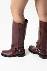 Botas de caña alta en cuero con textura de cocodrilo, color bordó. Presentan un diseño de punta cuadrada, taco bajo y una correa decorativa con argolla metálica y remaches en la zona del tobillo.