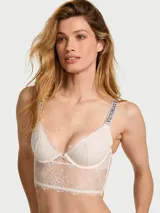 Bralette largo de encaje color blanco con aros, escote pronunciado en V, aberturas entrecruzadas al frente y tirantes brillantes con la marca Victoria's Secret en pedrería.