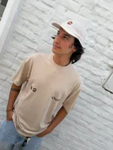 Remera oversize color beige con cuello redondo y manga corta. Presenta un estampado minimalista en color marrón en el pecho, que incluye un símbolo similar a un sol o estrella y texto.