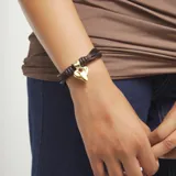Pulsera compuesta por múltiples hilos trenzados de efecto seda marrón y tiras de cuero negro, con un dije central metálico dorado con forma de corazón estilizado. Cuenta con cierres y argollas de metal dorado.