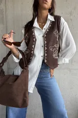 Chaleco color chocolate con bordados en hilo blanco en los bordes y frente. Presenta detalles de tachas metálicas en los hombros y cierre frontal.