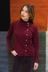 Remera de manga larga color bordó, con cuello alto y cierre frontal asimétrico con botones. Presenta detalles de volados en el cuello, en la línea de los botones y en los puños.