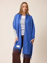 Pashmina de textura suave y color crema, con diseño liso y terminaciones en flecos en los extremos. Incluye una etiqueta decorativa en una de las esquinas.